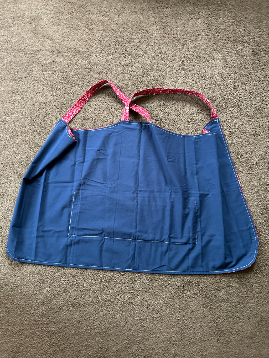 The item in the image is a cross-back kitchen apron (often referred to as a Japanese-style pinafore apron). It features a red patterned design with shoulder straps that cross over the back, along with a solid blue front utility pocket.