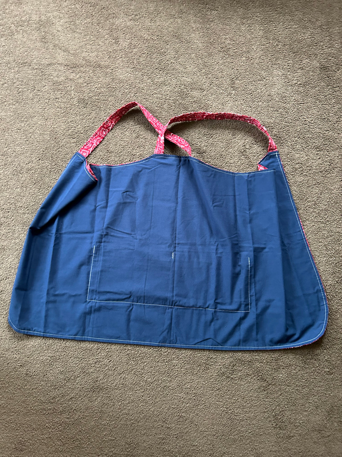 The item in the image is a cross-back kitchen apron (often referred to as a Japanese-style pinafore apron). It features a red patterned design with shoulder straps that cross over the back, along with a solid blue front utility pocket.