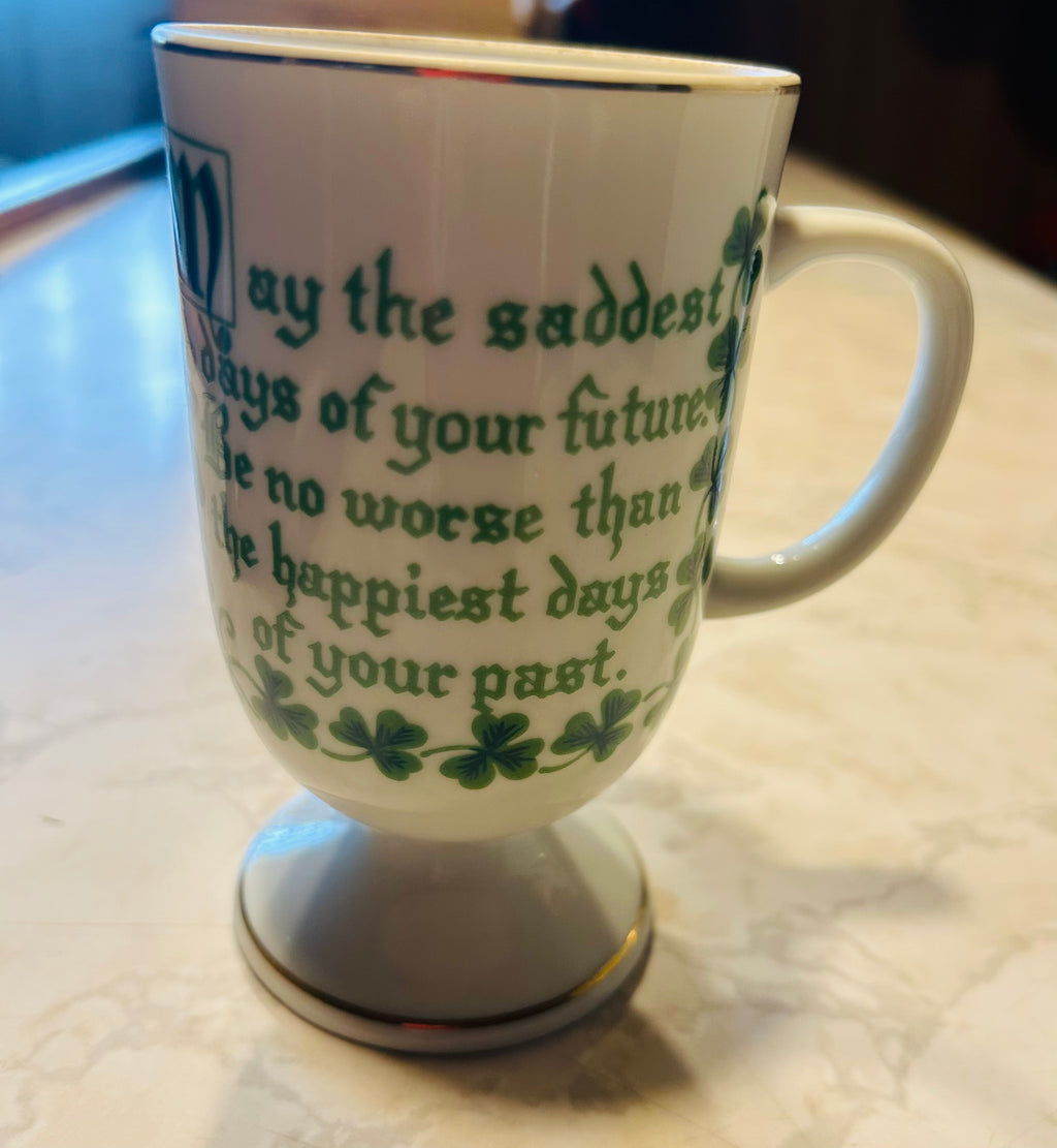 This is a white porcelain footed mug featuring green shamrocks, gold trim, and an Irish blessing.