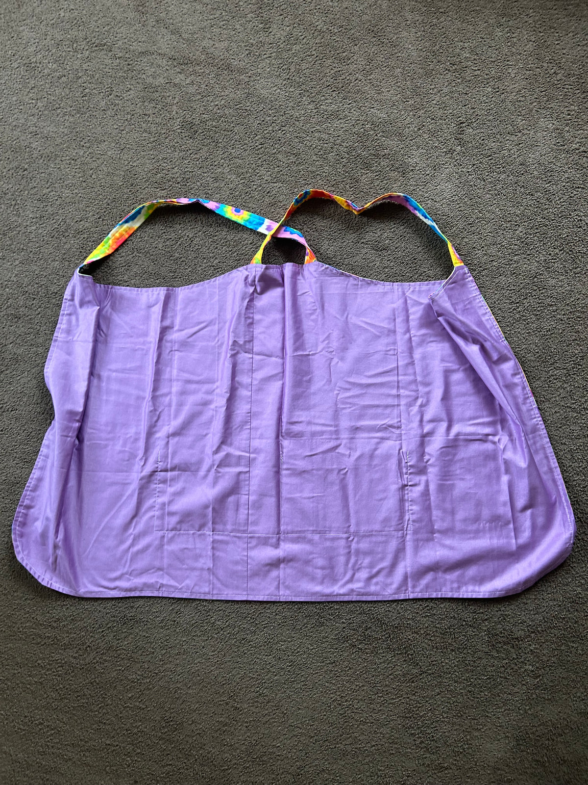 This item is a cross-back style kitchen apron designed without neck straps to distribute weight evenly across your shoulders. It features a vibrant, colorful tie-dye or flower-power circular print, a curved bottom hem, and a wide front pocket for holding