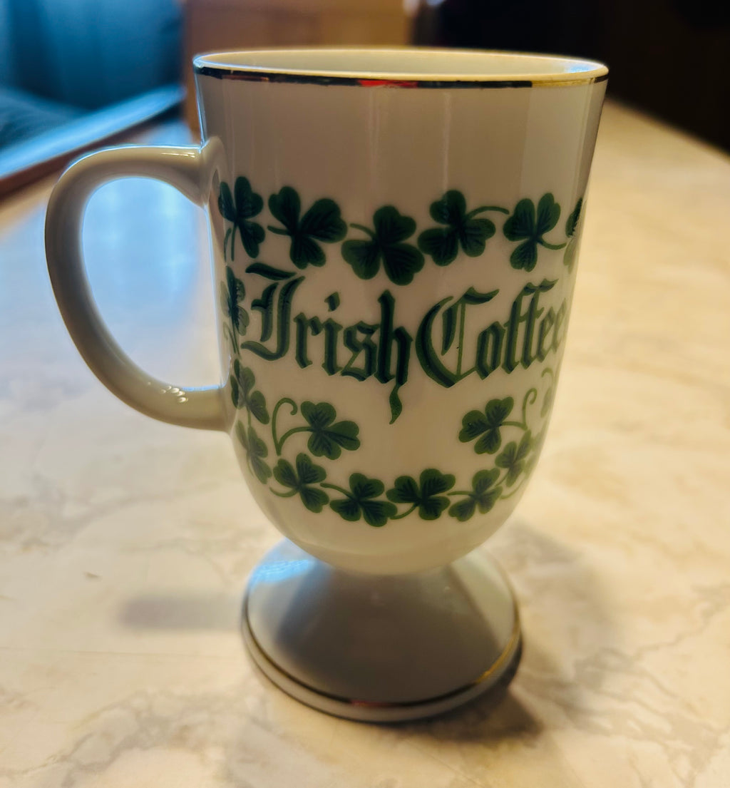 This is a white porcelain footed mug featuring green shamrocks, gold trim, and an Irish blessing.