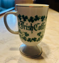 This is a white porcelain footed mug featuring green shamrocks, gold trim, and an Irish blessing.