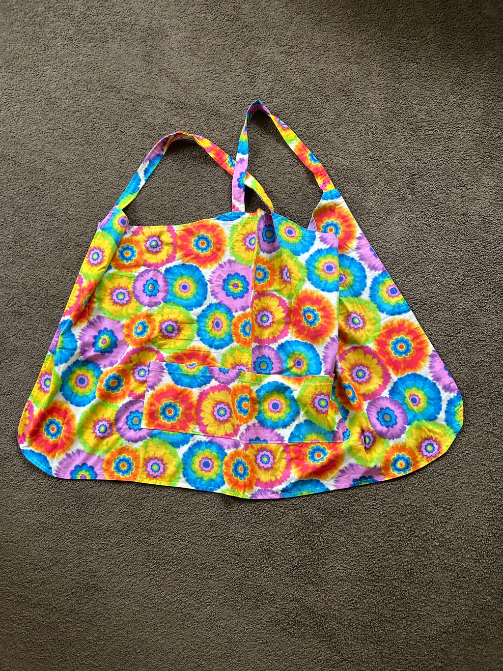 This item is a cross-back style kitchen apron designed without neck straps to distribute weight evenly across your shoulders. It features a vibrant, colorful tie-dye or flower-power circular print, a curved bottom hem, and a wide front pocket for holding