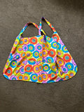 This item is a cross-back style kitchen apron designed without neck straps to distribute weight evenly across your shoulders. It features a vibrant, colorful tie-dye or flower-power circular print, a curved bottom hem, and a wide front pocket for holding
