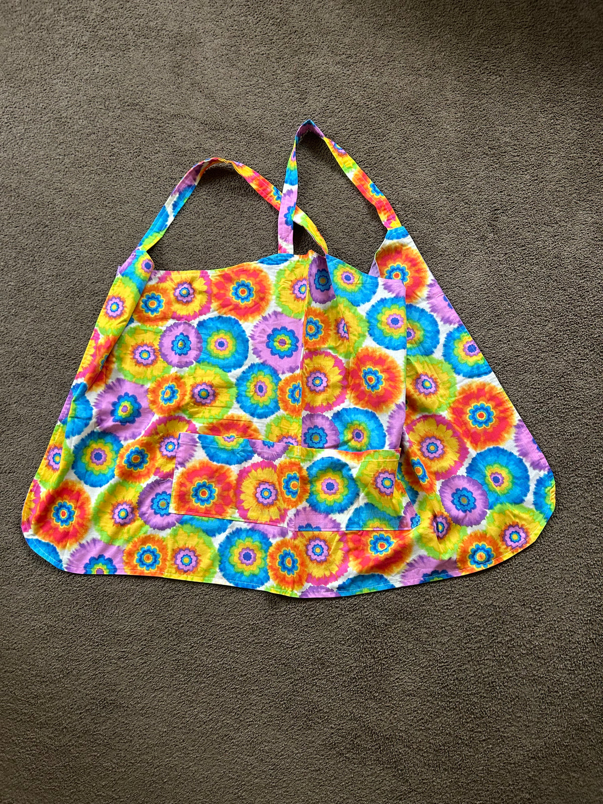 This item is a cross-back style kitchen apron designed without neck straps to distribute weight evenly across your shoulders. It features a vibrant, colorful tie-dye or flower-power circular print, a curved bottom hem, and a wide front pocket for holding