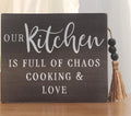 This is a black Parisloft wood tabletop sign with white text and a beaded jute tassel. “Our kitchen is full of chaos cooking & love.”